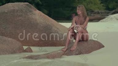 塞舌尔。 普拉斯林岛。 穿着泳装的黝黑美丽的女人，苗条的身体坐在蓝色的大石头上
